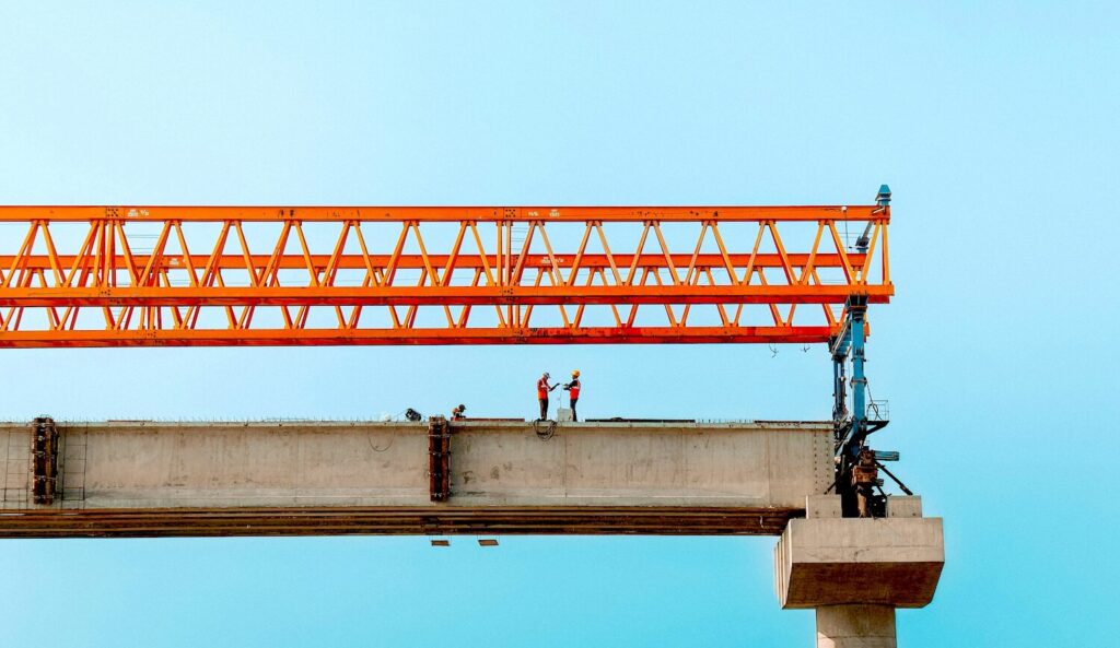 Construction workers standing on an elevated concrete bridge beam beneath an orange gantry crane, illustrating infrastructure work and construction wage trends.