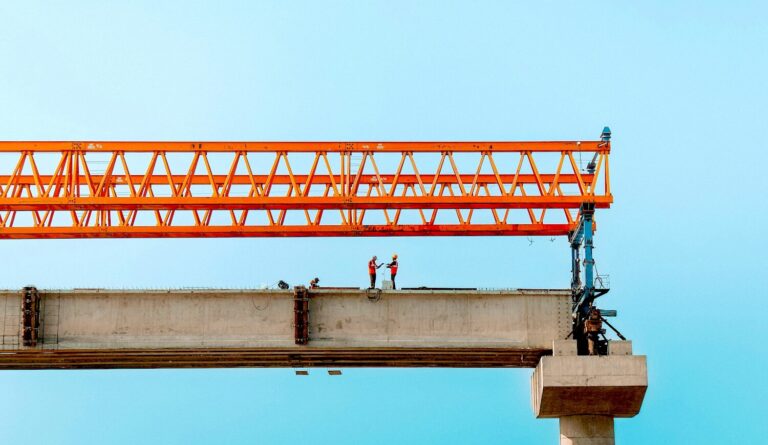 Construction workers standing on an elevated concrete bridge beam beneath an orange gantry crane, illustrating infrastructure work and construction wage trends.